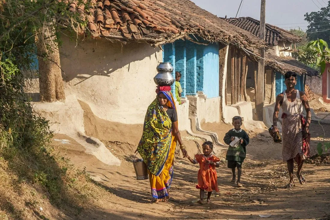 A woman carrying a pot on her head through a village lane