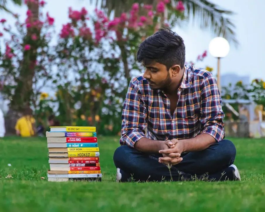 A young man sitting on the grass beside a stack of books